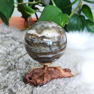 Pode incluir: Uma esfera de ágata polida e veteada sobre um suporte de madeira. A esfera tem um padrão em camadas de marrom, branco e cinza. O suporte é de madeira natural com uma textura áspera.