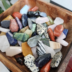 Op de afbeelding: Een verzameling kleine, doodskistvormige gepolijste edelstenen in diverse kleuren en patronen, waaronder zwarte, witte, rode, groene, blauwe en gevlekte variëteiten, allemaal in een vierkante houten kom met een zichtbare houtnerfstructuur.