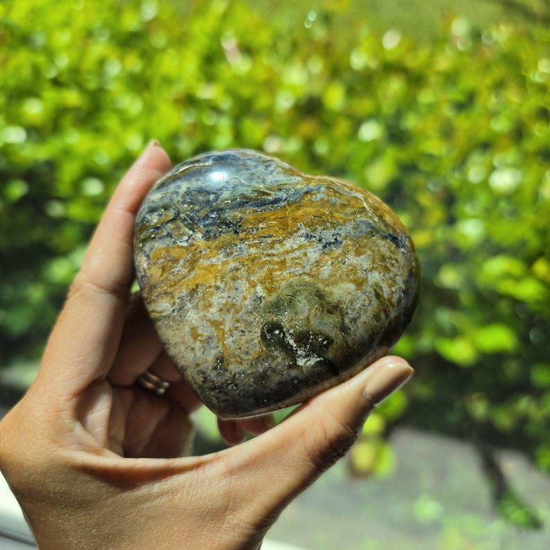 May include: A heart-shaped polished stone featuring a complex pattern of swirling colors including deep forest green, earthy brown, mustard yellow, and charcoal gray with a smooth, glossy finish and natural mineral inclusions throughout.