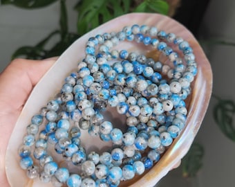 May include: A strand of blue and white kyanite beads, approximately 6mm in size, displayed on a shell. The beads have a speckled appearance with a mix of light blue and white.