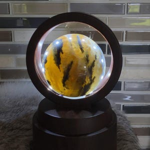 May include: A spherical yellow and black patterned stone orb displayed within a circular wooden frame. The orb is illuminated from behind by integrated LED lights, highlighting the natural dark inclusions and golden hues of the mineral sphere.