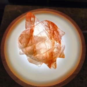 Puede incluir: Un cristal transparente y facetado con estrías naranjas y rojas. El cristal tiene forma de estrella y está iluminado desde abajo.