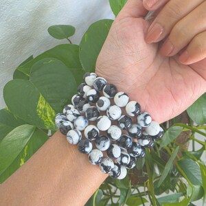 Op de afbeelding: Een elastische armband bestaande uit meerdere ronde kralen met een gemarmerd patroon in zwart en wit. De kralen zijn gerangschikt in een ontwerp met meerdere strengen, wat zorgt voor een samenhangende uitstraling rond de pols met een gladde afwerking.