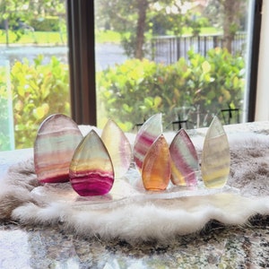 May include: A collection of seven iridescent fluorite crystals, each with a unique color pattern of pink, green, yellow, and white, arranged on a white, fluffy surface.
