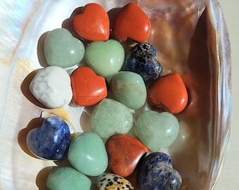 Könnte beinhalten: Eine Sammlung herzförmiger Steine in verschiedenen Farben, darunter Rot, Grün, Weiß und Blau, eingebettet in einer perlmuttartigen Muschelschale. Die Steine variieren in Textur und Muster und bilden eine ansprechende Darstellung.