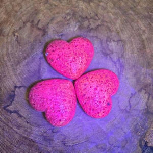 May include: Three pink heart-shaped stones with a textured surface, arranged in a triangular pattern on a wooden background.