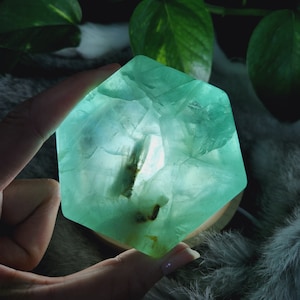 May include: A green fluorite crystal with a geometric shape, illuminated from within, creating a translucent effect. The crystal is resting on a wooden base.