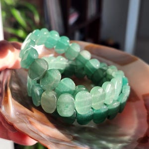Puede incluir: Se muestran dos pulseras de aventurina verde, cada una compuesta por cuentas ovaladas. Las pulseras descansan en una concha, mostrando el color verde natural de las piedras. Las cuentas tienen un acabado pulido.