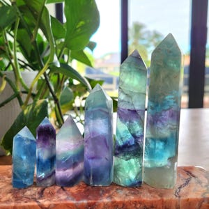 Könnte beinhalten: Ein Satz von sieben Fluorit-Kristallspitzen in verschiedenen Blautönen, Grüntönen und Violetttönen. Die Kristalle sind in einer Reihe vom kleinsten zum größten angeordnet.