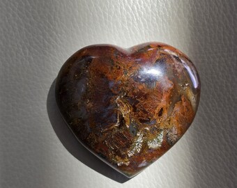 May include: A polished heart-shaped stone featuring a complex pattern of deep brown, rust, and amber tones with translucent areas. The surface is smooth and reflective, showcasing natural mineral inclusions and varied earthy color gradients.