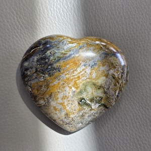 May include: A polished heart-shaped stone featuring a complex marbled pattern with swirling colors of deep ochre, muted olive green, charcoal gray, and creamy white. The surface is smooth and reflective, showcasing natural mineral inclusions.