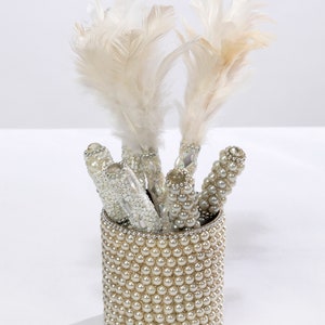 Könnte beinhalten: Ein weißer, mit Perlen bedeckter zylindrischer Behälter mit einem Satz weißer Staubwedel.