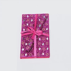 May include: A magenta notebook with a decorative cover featuring small mirrors, gold circles, and diamond accents. A matching magenta ribbon is tied in a bow around the notebook.