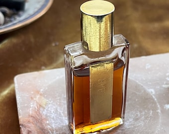 Può includere: Un flacone di profumo rettangolare in vetro riempito con liquido ambrato, dotato di un tappo cilindrico color oro e un'etichetta verticale dorata sul davanti con il testo ESTEE LAUDER CINNABAR stampato in caratteri chiari.