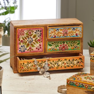 以下が含まれることがあります： カラフルな花柄が描かれた引き出しが4つ付いた木製のジュエリーボックス。マンゴーウッド製で、ナチュラルな仕上げ。引き出しには、手の込んだ手描きのディテールが施されています。