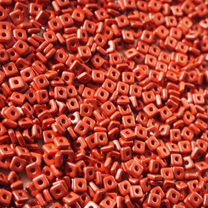 Puede incluir: Una gran cantidad de pequeñas cuentas cuadradas rojas con un acabado metálico. Las cuentas tienen un agujero en el centro para enhebrar.