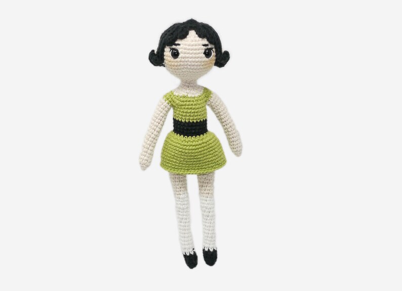 May include: A crocheted doll with black hair, green dress, and white legs. The doll has a black belt and black shoes.