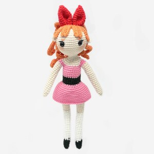 May include: A crocheted doll with orange hair, a pink dress, and a red bow. The doll has black eyes and a white face.