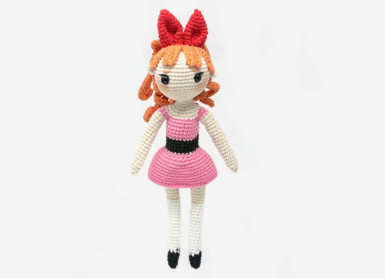 May include: A crocheted doll with orange hair, a pink dress, and a red bow. The doll has black eyes and a white face.