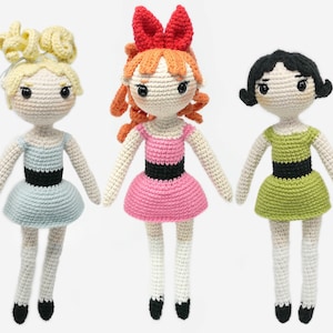 May include: Three crocheted dolls with different colored dresses and hair. The doll on the left has blonde hair and a light blue dress. The doll in the middle has orange hair and a pink dress. The doll on the right has black hair and a green dress.