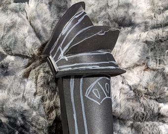 May include: Black foam armor piece with white lines and shapes drawn on it. The armor is shaped like a leg and foot.