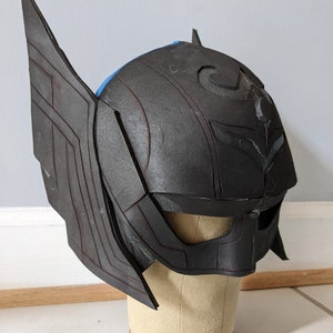 May include: A black helmet with a detailed design, including wings on the sides and a stylised symbol on the front. The helmet is made of foam and is displayed on a mannequin head.