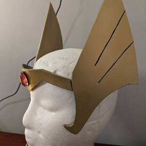 May include: A gold headband with two large, pointed wings on either side. The headband has a red gem in the center.