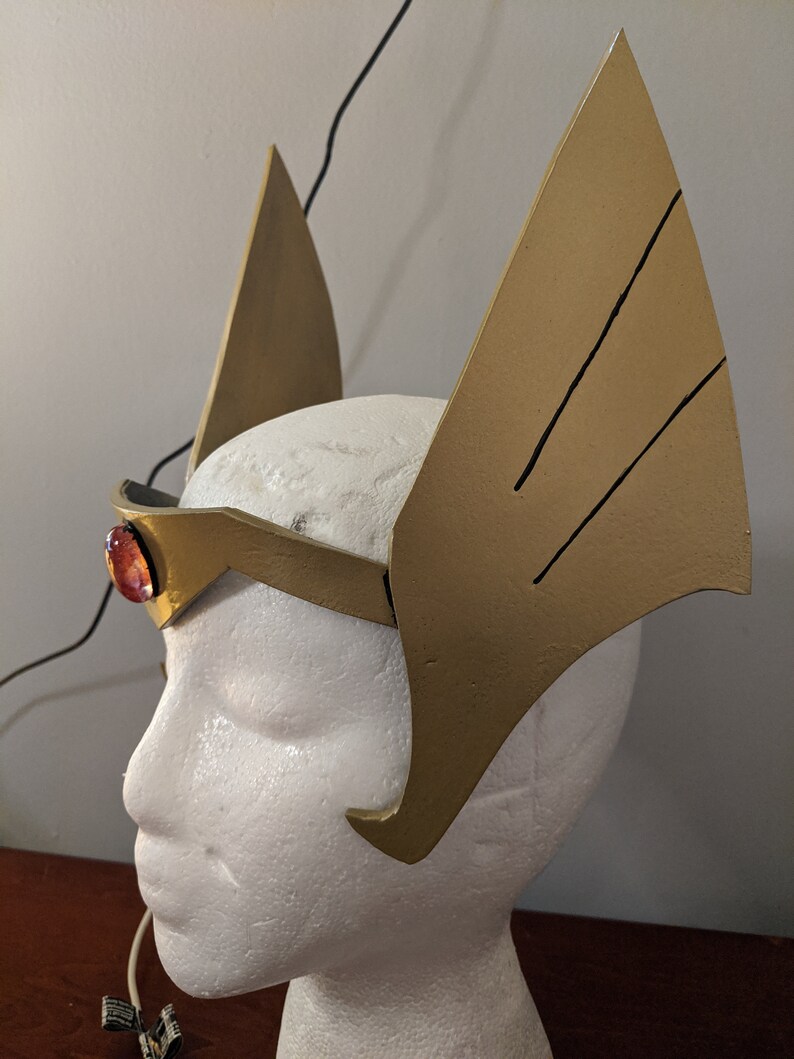 May include: A gold headband with two large, pointed wings on either side. The headband has a red gem in the center.
