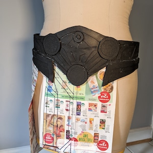 May include: A black, geometric belt design, possibly for a costume or cosplay, is displayed on a mannequin. The belt features circular and angular shapes. The mannequin is covered with a newspaper-like material.