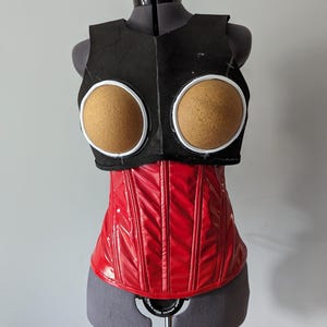 May include: A red corset with a black fabric top and two brown foam circles. The corset is on a dress form.