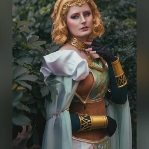 May include: A woman in a detailed cosplay costume with a gold headpiece, choker, and arm cuffs. The costume features a green and brown bodice, white sleeves, and a flowing cape. The image is set outdoors with greenery in the background. Text at the bottom reads "MAGFEST 2024 - ALEXANDRA LEE STUDIOS."