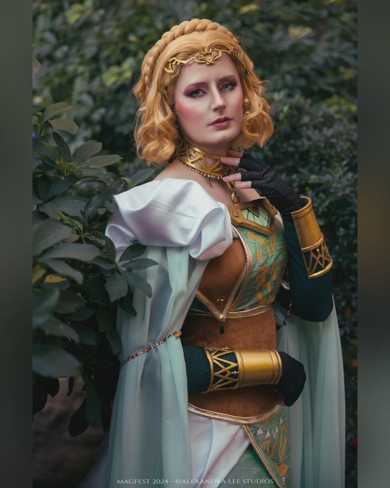 May include: A woman in a detailed cosplay costume with a gold headpiece, choker, and arm cuffs. The costume features a green and brown bodice, white sleeves, and a flowing cape. The image is set outdoors with greenery in the background. Text at the bottom reads "MAGFEST 2024 - ALEXANDRA LEE STUDIOS."