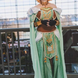 May include: A cosplayer in a detailed fantasy-inspired costume. The outfit features a mint green dress with gold accents, a brown bodice, and white accents. The cosplayer wears a gold necklace and headpiece, with black gloves. The background is a modern building.