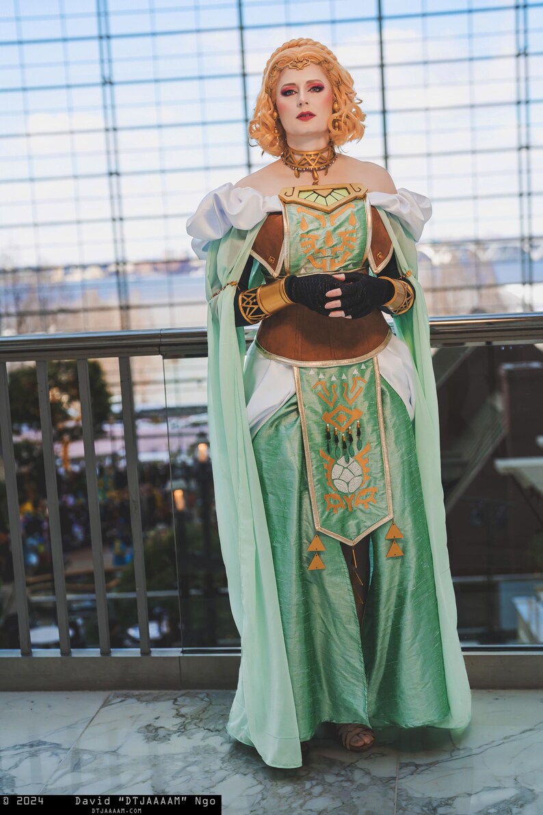May include: A cosplayer in a detailed fantasy-inspired costume. The outfit features a mint green dress with gold accents, a brown bodice, and white accents. The cosplayer wears a gold necklace and headpiece, with black gloves. The background is a modern building.