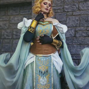 May include: A cosplay costume featuring a light blue and brown dress with gold accents. The dress has a detailed design on the front and is paired with a matching headpiece and arm cuffs. The costume is inspired by a fantasy theme.