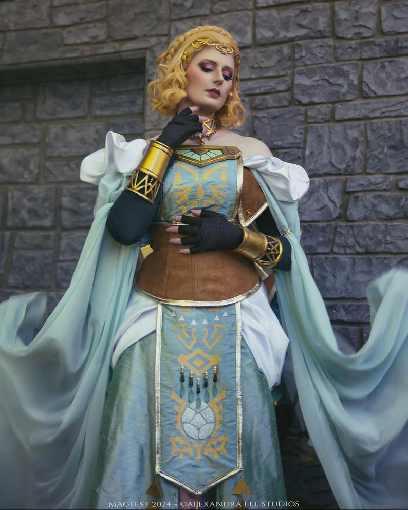 May include: A cosplay costume featuring a light blue and brown dress with gold accents. The dress has a detailed design on the front and is paired with a matching headpiece and arm cuffs. The costume is inspired by a fantasy theme.