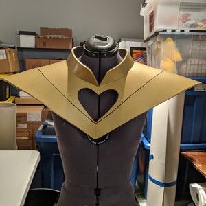 May include: A gold-colored, heart-shaped collar with a black outline. The collar is on a dress form.