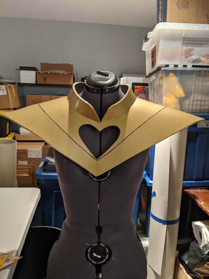 May include: A gold-colored, heart-shaped collar with a black outline. The collar is on a dress form.