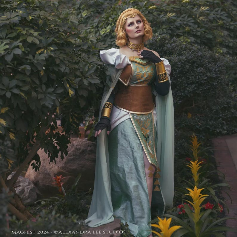 May include: A woman in an elaborate fantasy costume with a gold headpiece, choker, and armbands. The outfit features a teal and gold bodice, a brown corset, and a flowing teal skirt and cape. The image is set outdoors with lush greenery.
