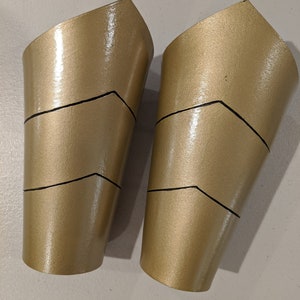 May include: Two gold-colored, plastic arm cuffs with black lines. The cuffs are shaped like a curved cylinder and have a pointed top.