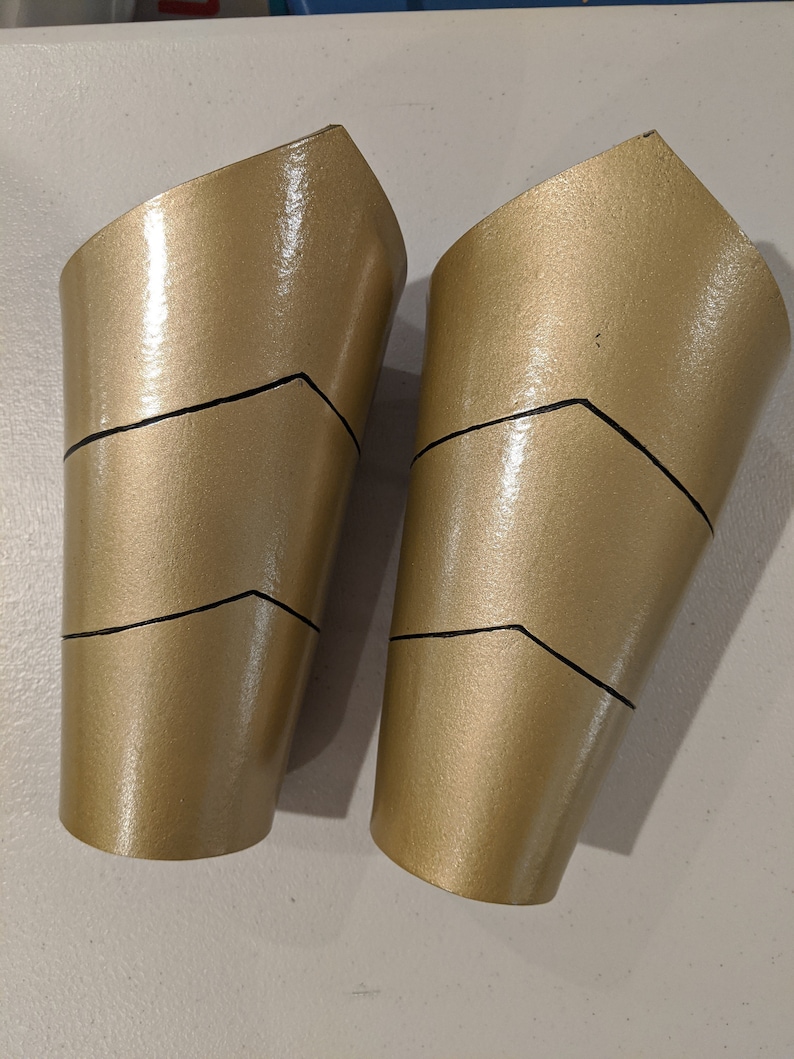 May include: Two gold-colored, plastic arm cuffs with black lines. The cuffs are shaped like a curved cylinder and have a pointed top.
