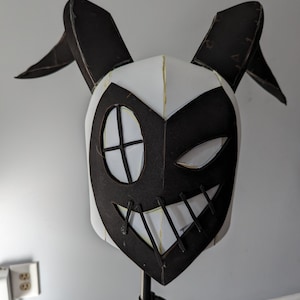 Puede incluir: Una máscara de papel maché blanca y negra con cuernos, un ojo grande y una sonrisa cosida. La máscara está montada en un soporte negro.