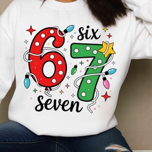 Peut inclure: Sweat-shirt blanc avec un motif festif représentant les chiffres "67" en rouge et vert, ornés de pois et de guirlandes lumineuses. Le mot "Six" est au-dessus du chiffre 6, et "Seven" est en dessous. Des étoiles et des éléments décoratifs complètent le design.