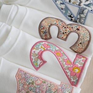 May include: Four white shirts with pink and floral fabric numbers sewn on them. The numbers are 1, 2, 3, and 4.