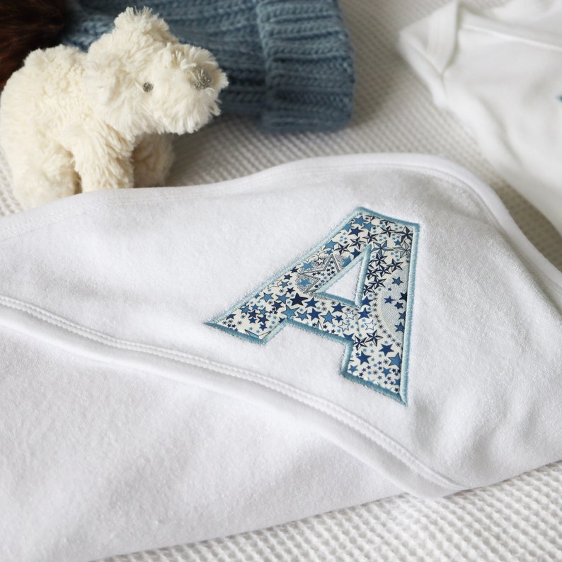 May include: A white baby blanket featuring a large embroidered letter "A" in a blue star design. A blue knitted hat and a white teddy bear are in the background. The blanket is folded, appearing soft and cosy.