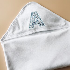 May include: White hooded baby towel featuring an embroidered letter "A" in blue and white star patterns. The towel is made of soft, absorbent material. The hood is visible, along with a portion of the folded towel, ideal for after bath time.