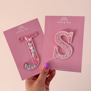 May include: Two pink cards featuring the letters "J" and "S" appliquéd with a floral fabric and white stitching. The cards display the text "Ollie & Eve" and a rainbow logo. The letters are for decorative purposes.