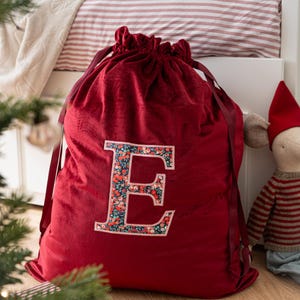 May include: A deep red velvet drawstring sack featuring a large, central letter E. The letter is filled with a dark blue floral pattern containing small red, white, and green flowers. The sack is cinched at the top with matching red ribbon ties.