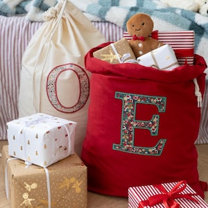 May include: A large red fabric sack featuring a prominent letter E with a floral pattern, sitting next to a cream-coloured sack with a letter O. The red sack is filled with wrapped gifts and a plush gingerbread person, with more gifts placed around it.