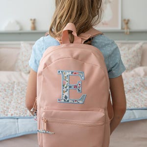 May include: A pale pink backpack featuring a floral embroidered letter 'E'. The backpack has a front zip pocket and adjustable straps. The girl is wearing a blue top and a floral bow in her hair.